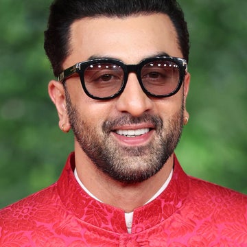 Man in a patterned jacket and sunglasses smiling in front of a leafy backdrop