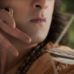 Person drawing a bow with focused expression, wearing beaded necklace and distinctive ring