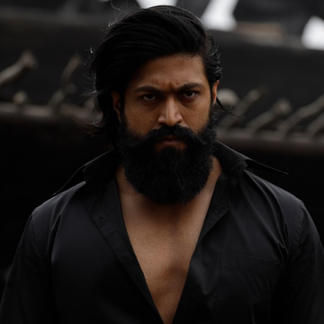 Person with long hair and beard wearing an open-collared black shirt, looking intensely at the camera against a blurred background