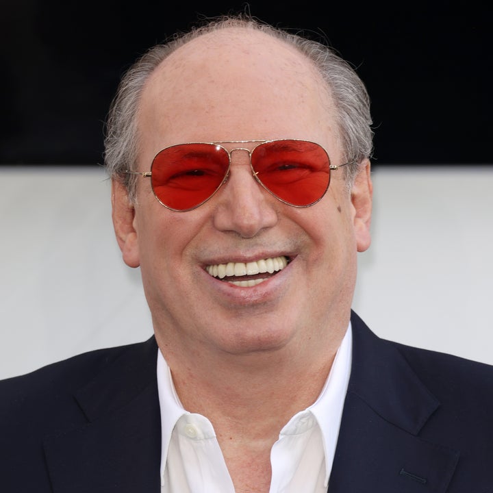 Smiling person wearing a dark suit, white shirt, and red-tinted sunglasses