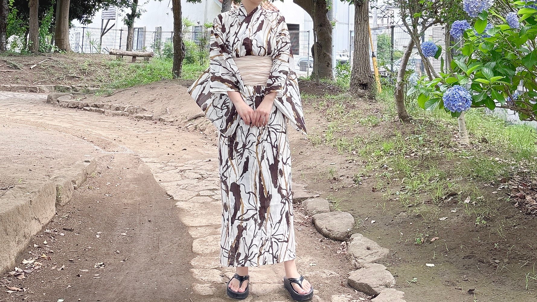 女性が伝統的な着物とサンダルを着用し、自然の中の小道に立っている。