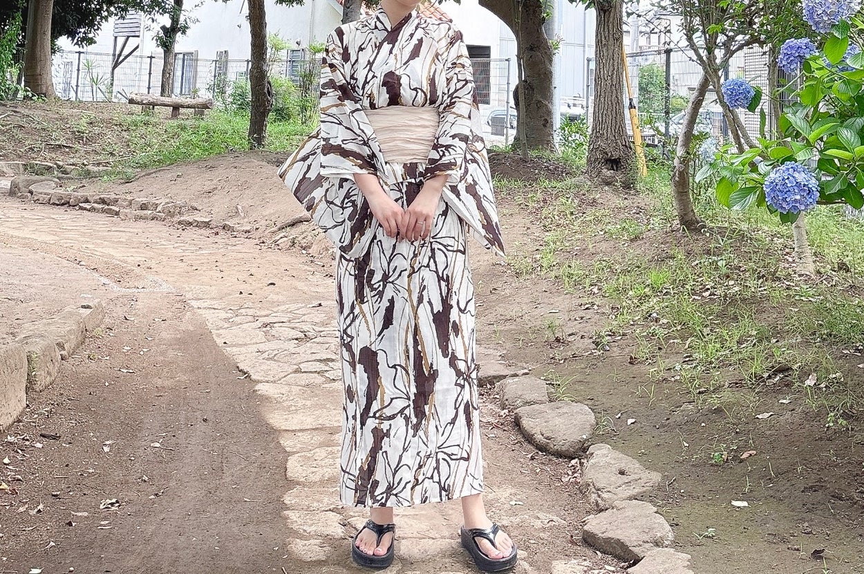 浴衣を着た人物が庭園を散策している様子。着物は木の枝の模様で、足元は草履を履いている。