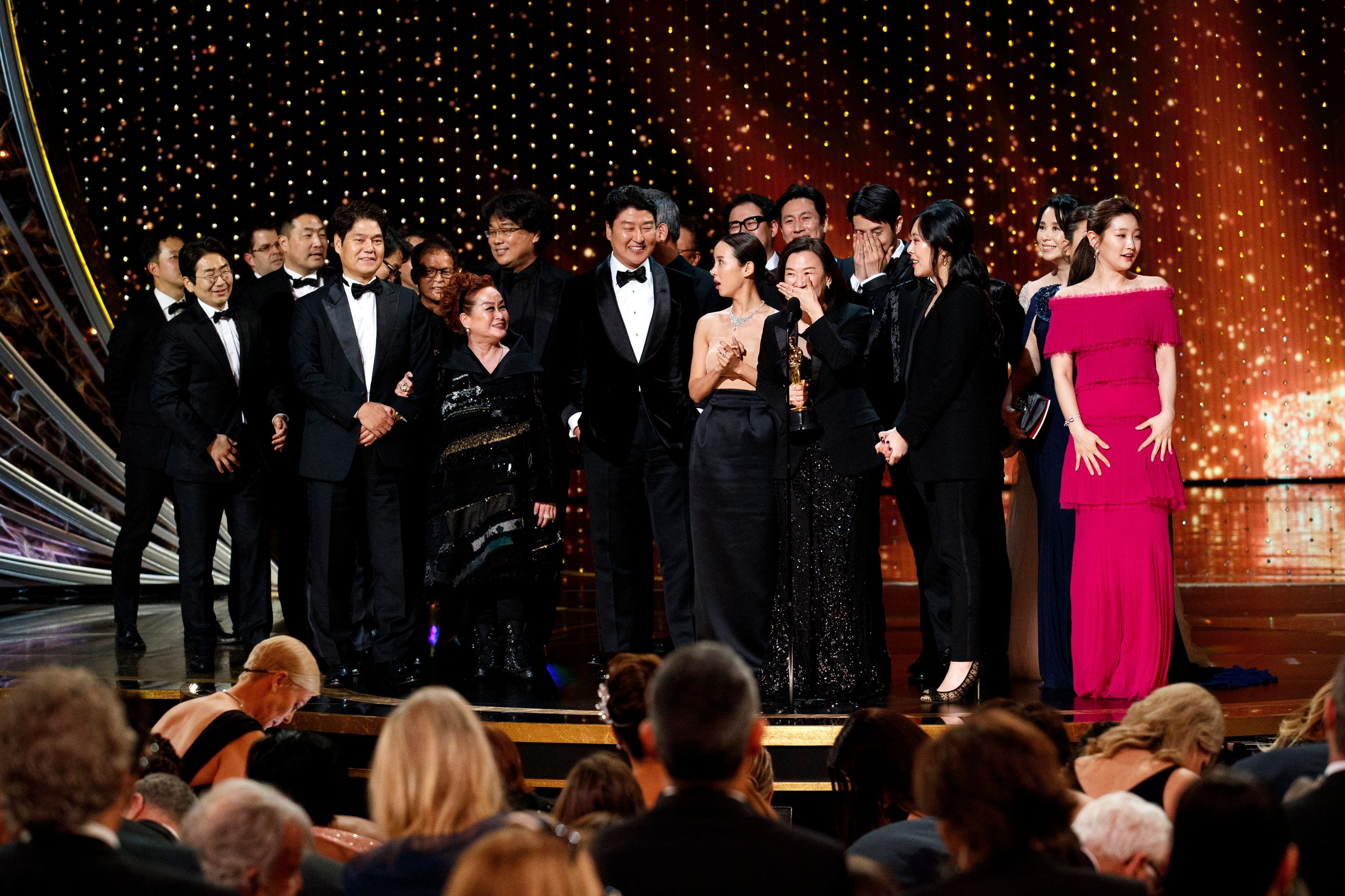 Group of people in formal attire on stage at an awards show, some holding trophies, celebrating an apparent victory. Audience is visible in foreground