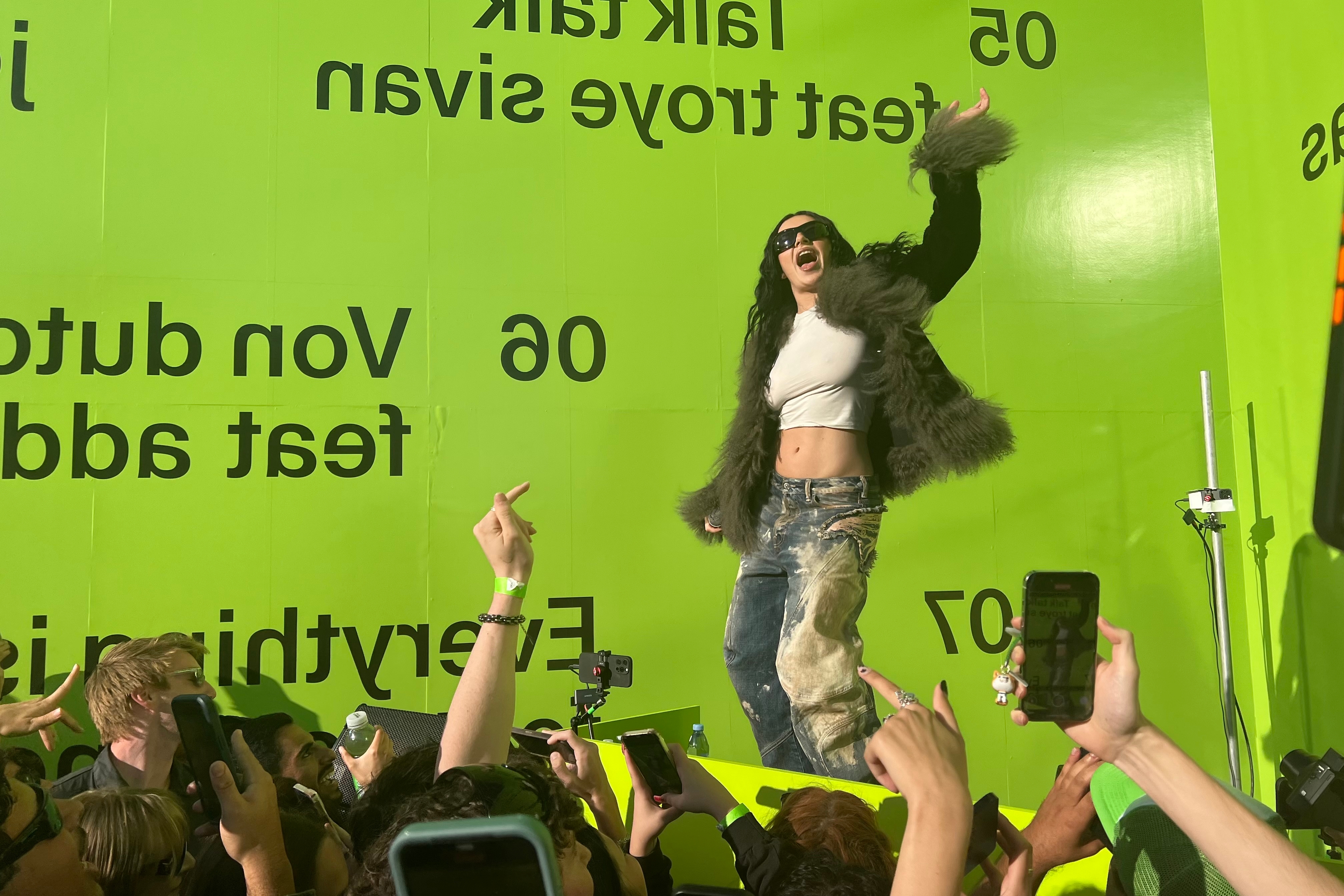 A person energetically performs on stage, wearing a furry jacket and distressed jeans, surrounded by an enthusiastic crowd