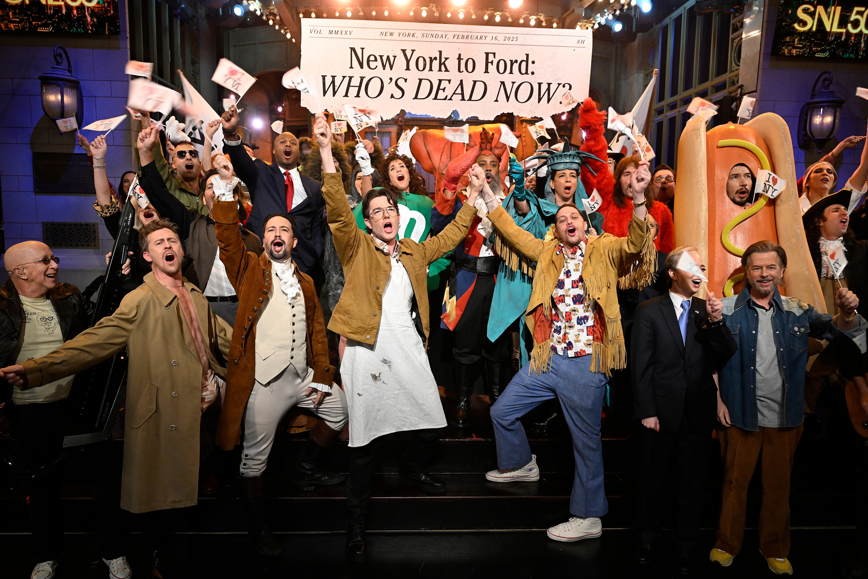 A group of people in theatrical costumes energetically pose on a stage under a sign reading "New York to Ford: Who's Dead Now?" during a live show