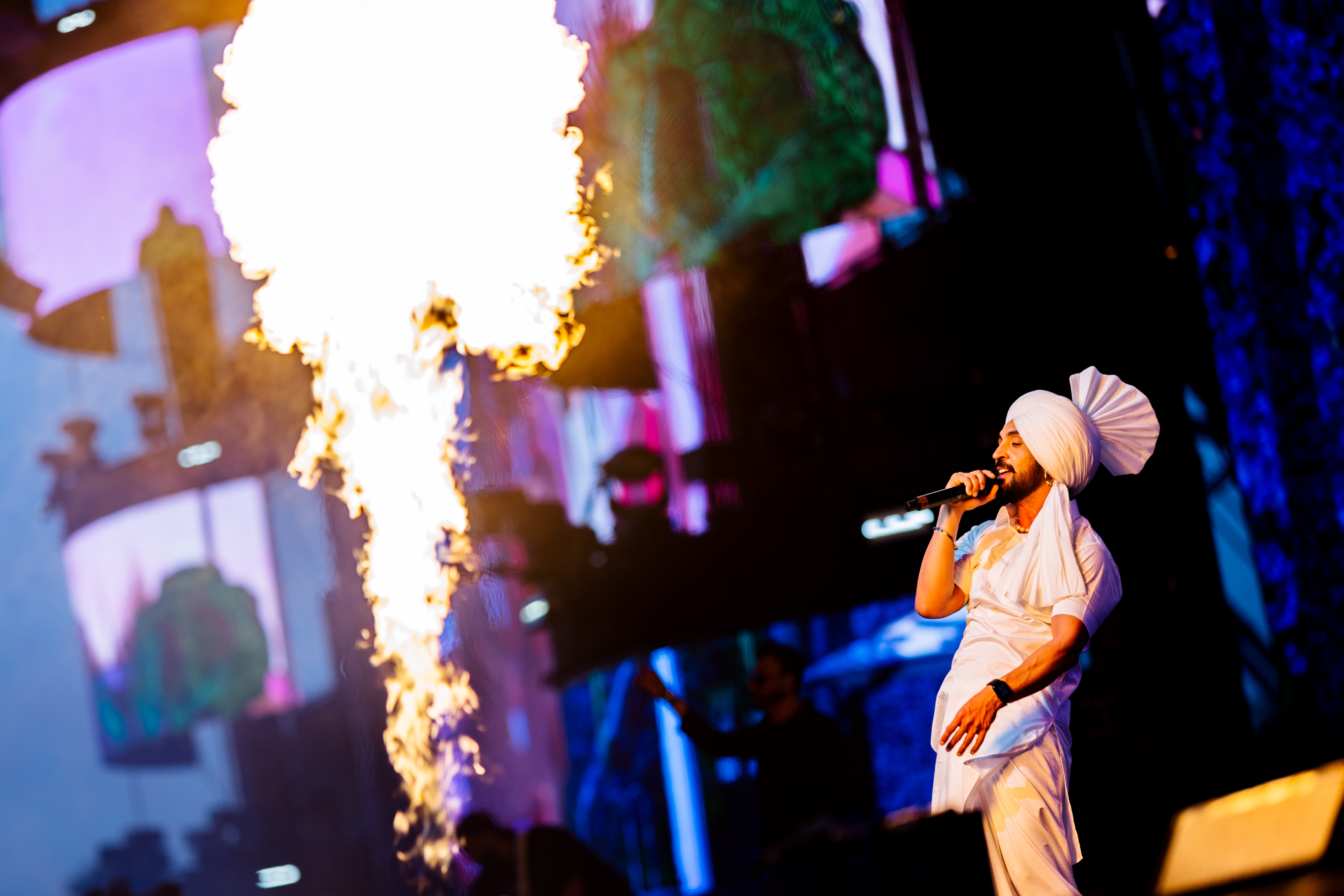 Performer in traditional attire sings on stage with large flame effect beside them