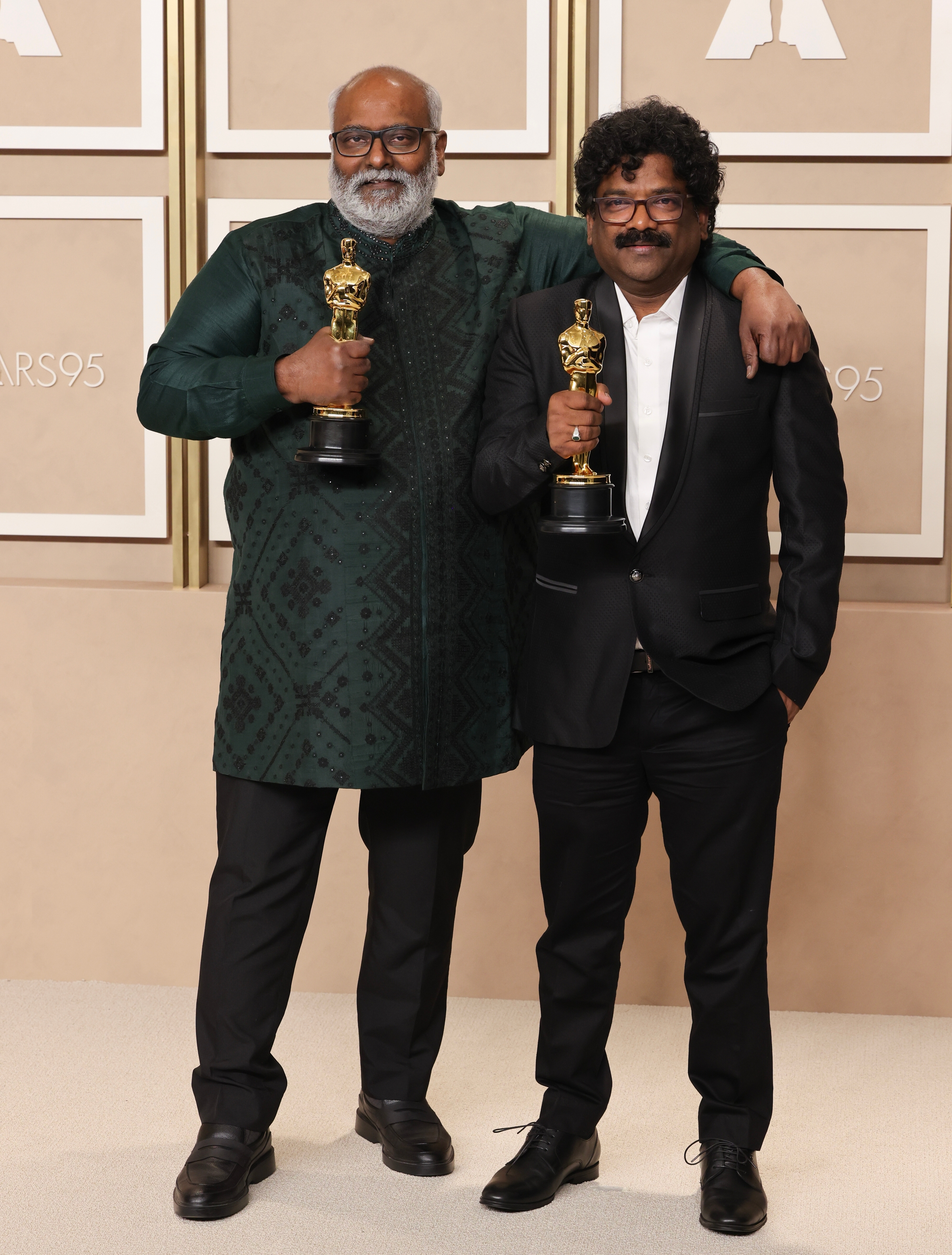 Two individuals pose with awards. One wears a patterned tunic; the other is in a suit. They stand on a stage with "Oscars 95" signage