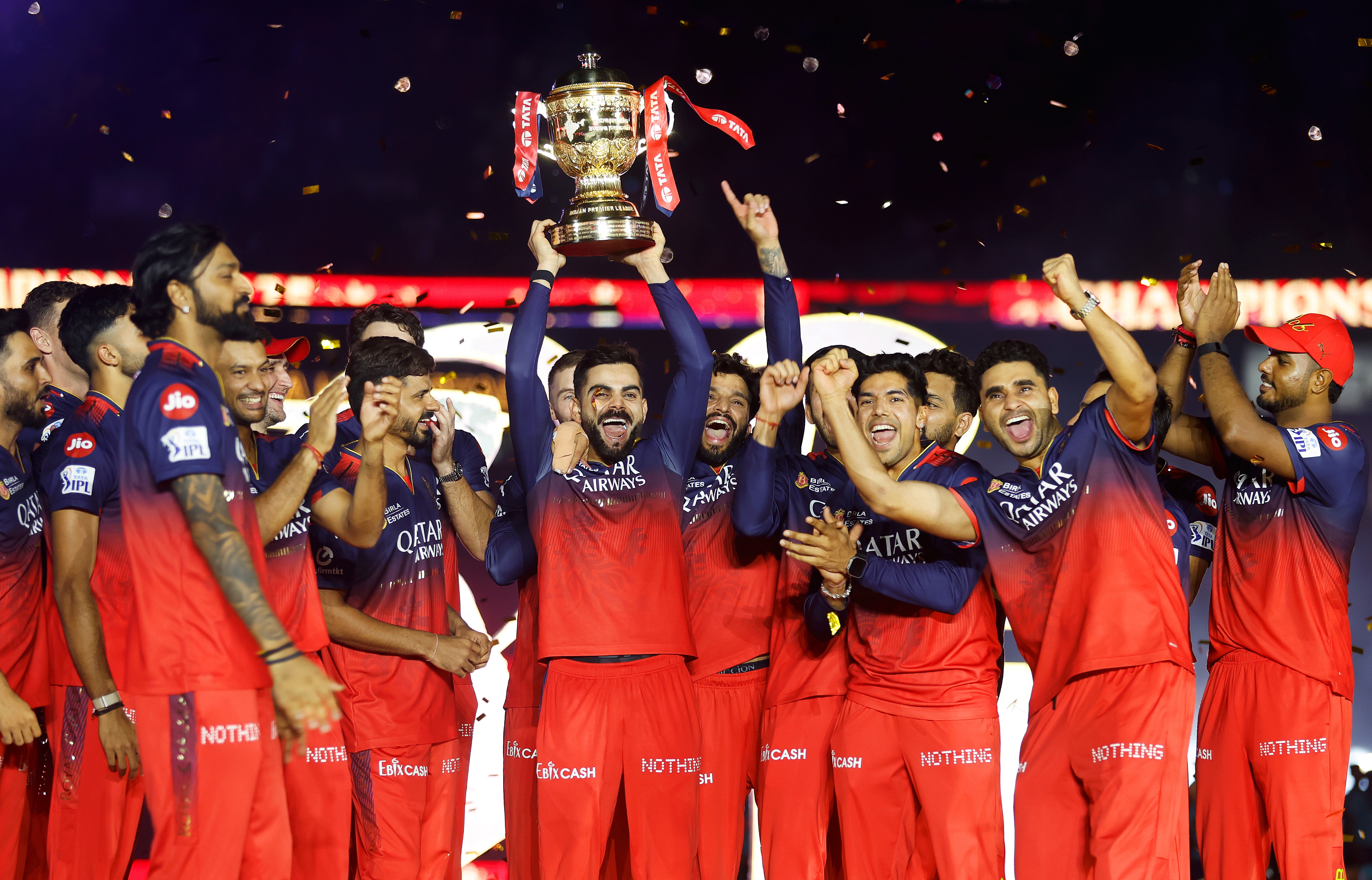 A cricket team in matching sporty attire celebrates, holding up a large trophy with joy and enthusiasm
