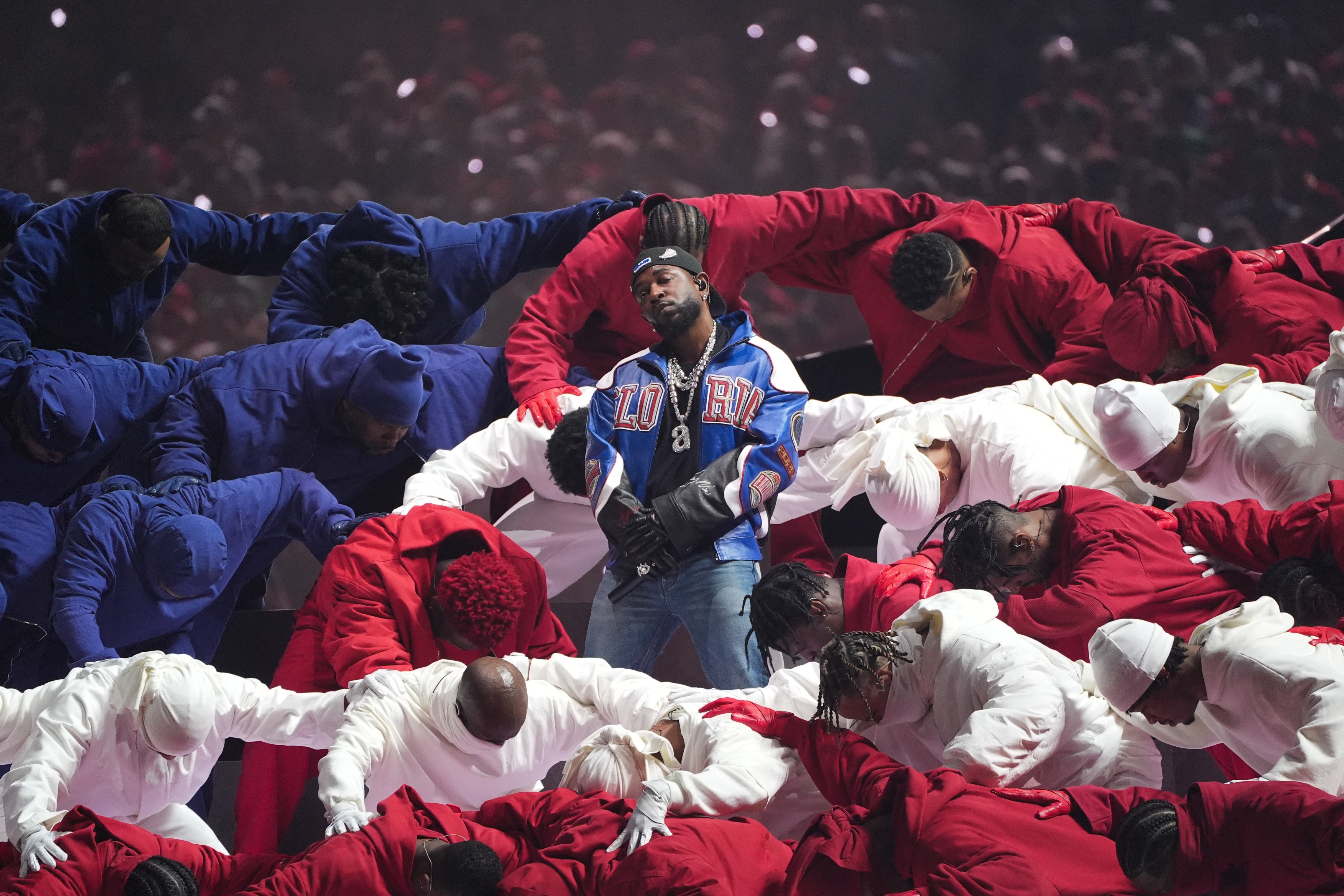 A performer in a blue sports jacket stands center stage, surrounded by dancers in red and white hoodies bending towards him