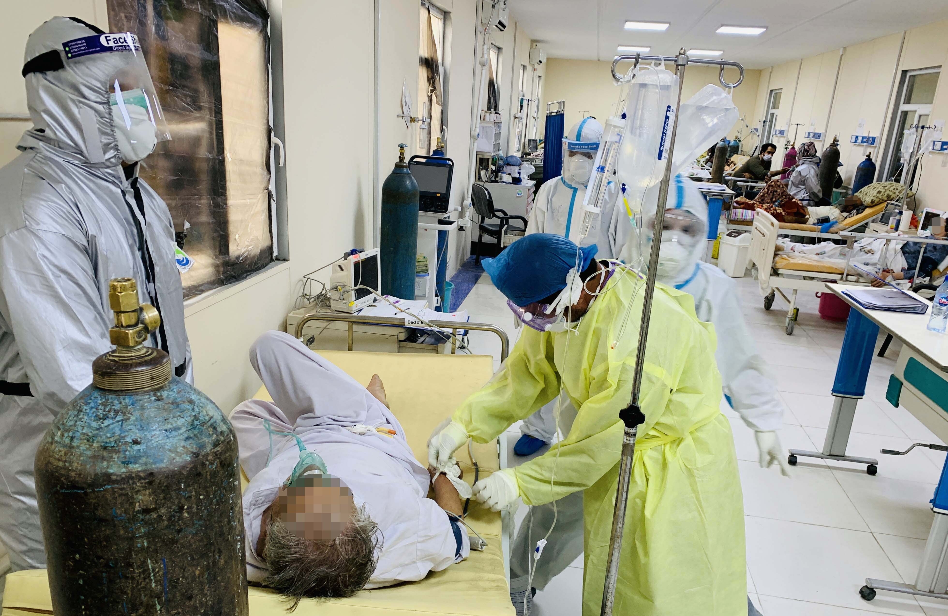 Healthcare workers in protective gear attend to patients in a busy hospital ward. One patient lies on a bed receiving care