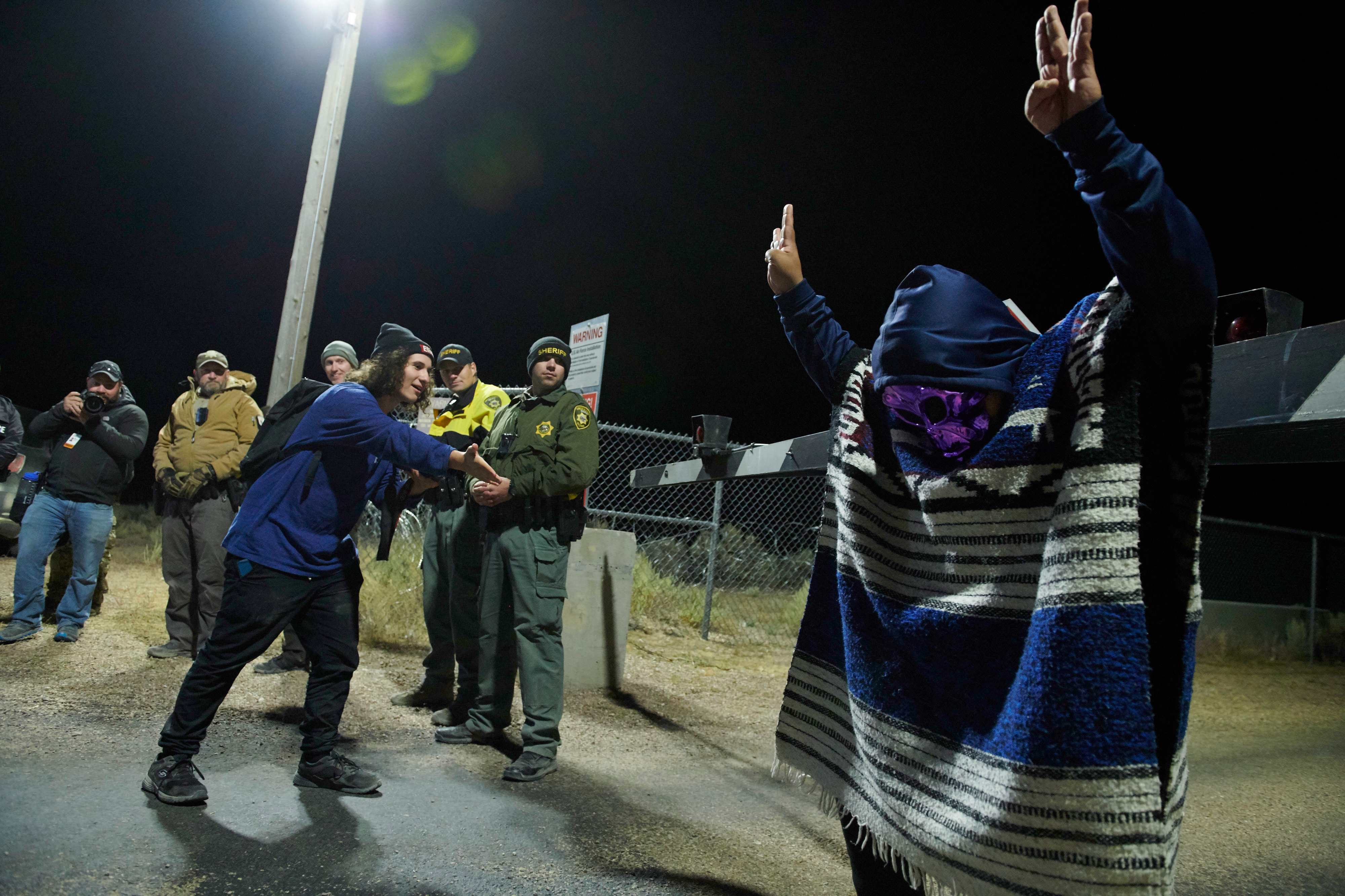 Person in blanket raises hands near guardrail as others observe near fenced area at night