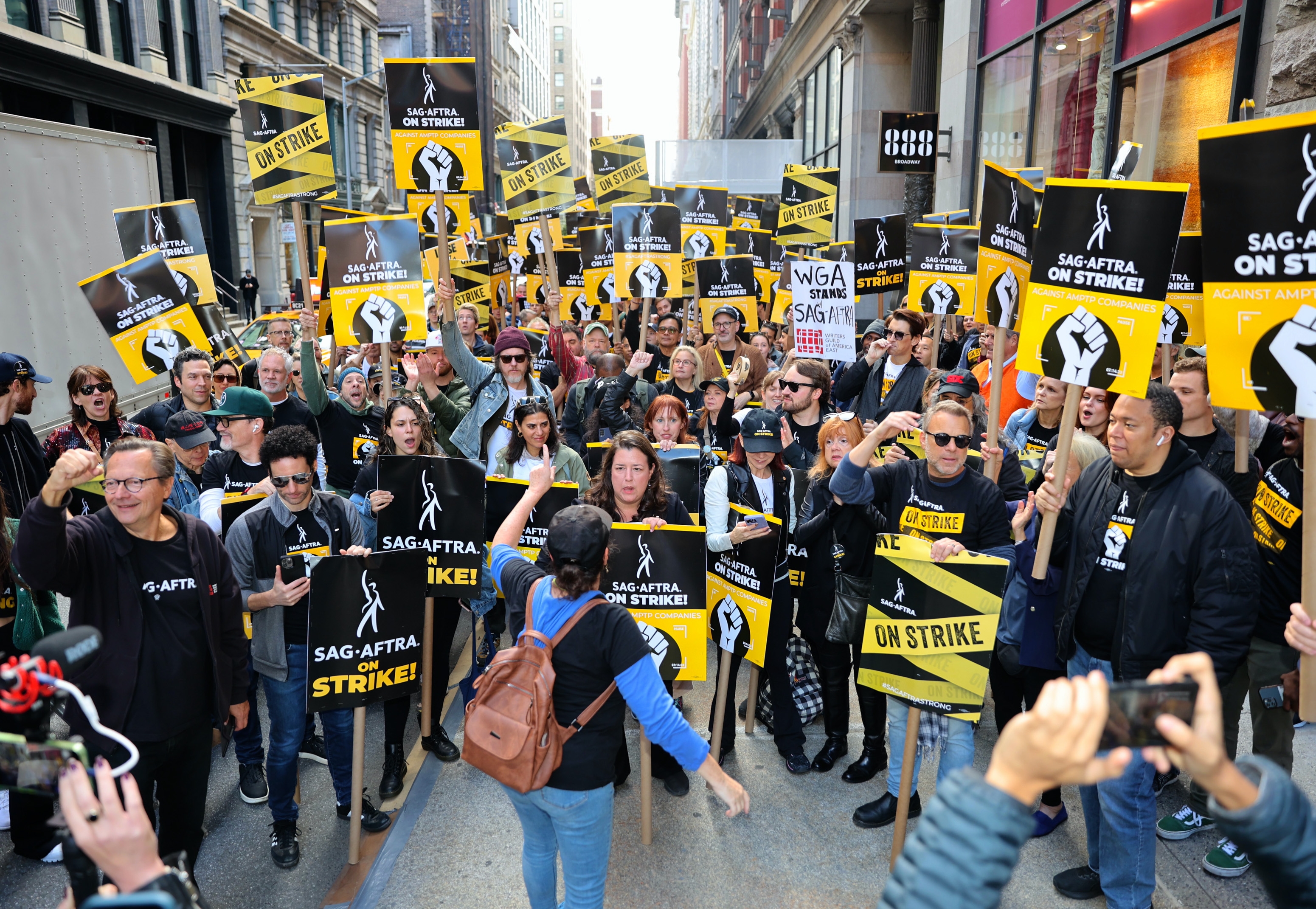 A large crowd of people hold signs saying "SAG-AFTRA On Strike" during a street protest, with a few photographers capturing the scene