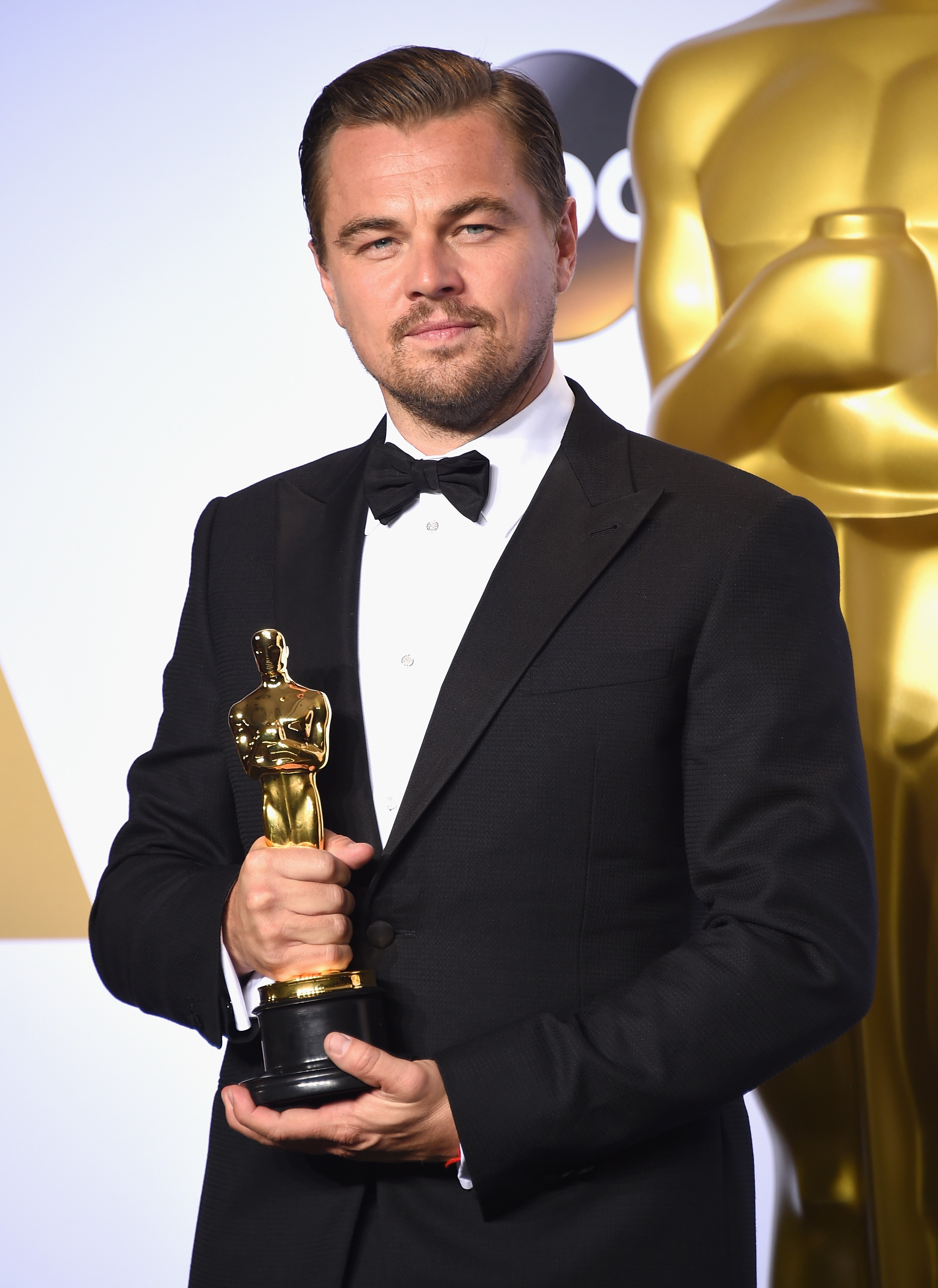 Person in a tuxedo holding an Oscar statue, standing in front of a large Oscar award statuette