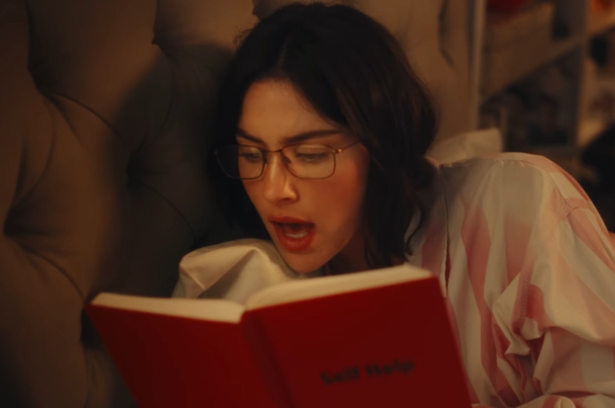 Gracie Abrams in a music video in pajamas and glasses lying in bed, engrossed in reading a red book, with an expression of surprise or intrigue