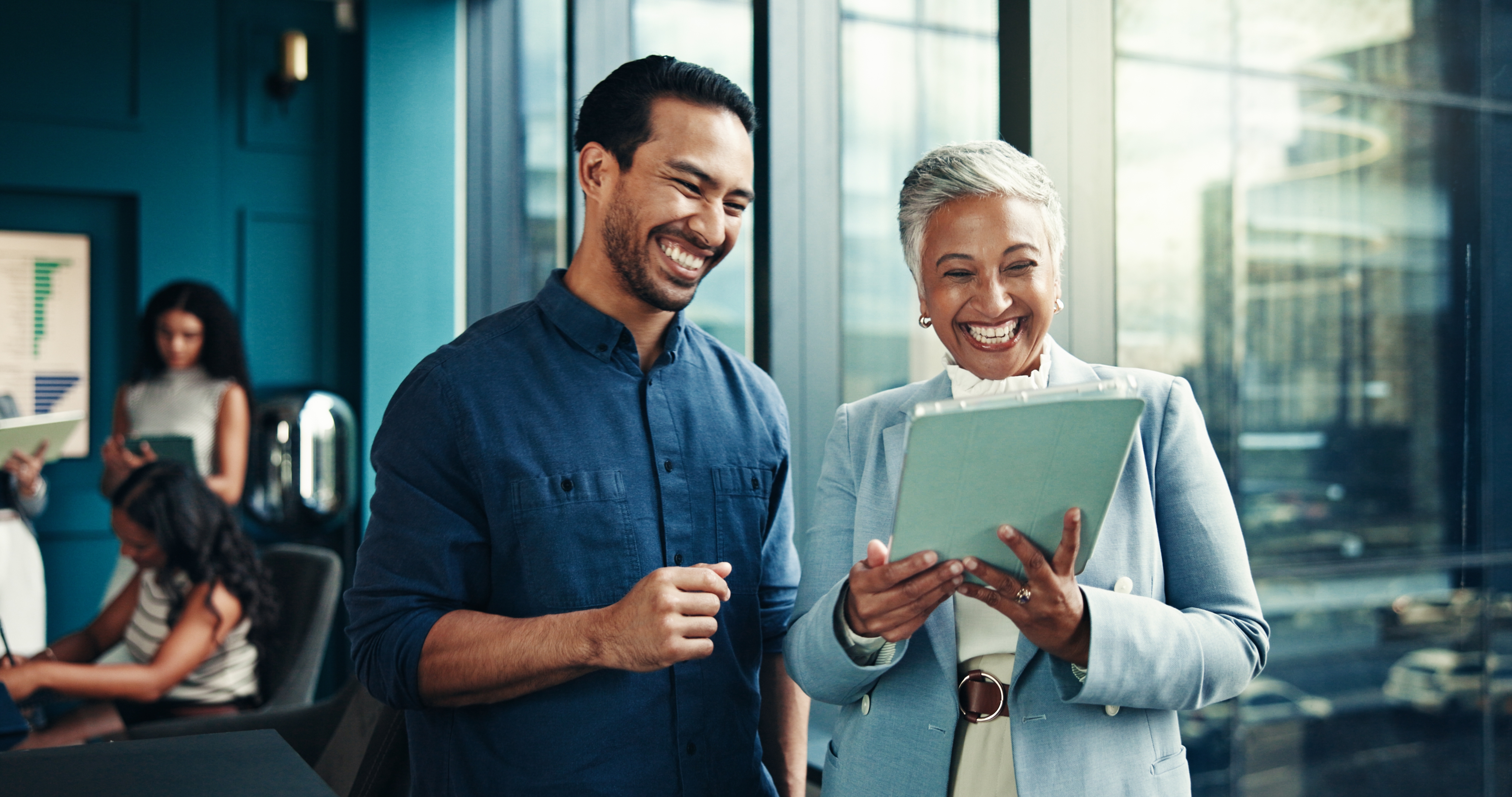 To personer som smiler mens de ser på et nettbrett i et kontorinnstilling. En kvinne i dress og en mann i en tilfeldig skjorte er engasjert i en diskusjon
