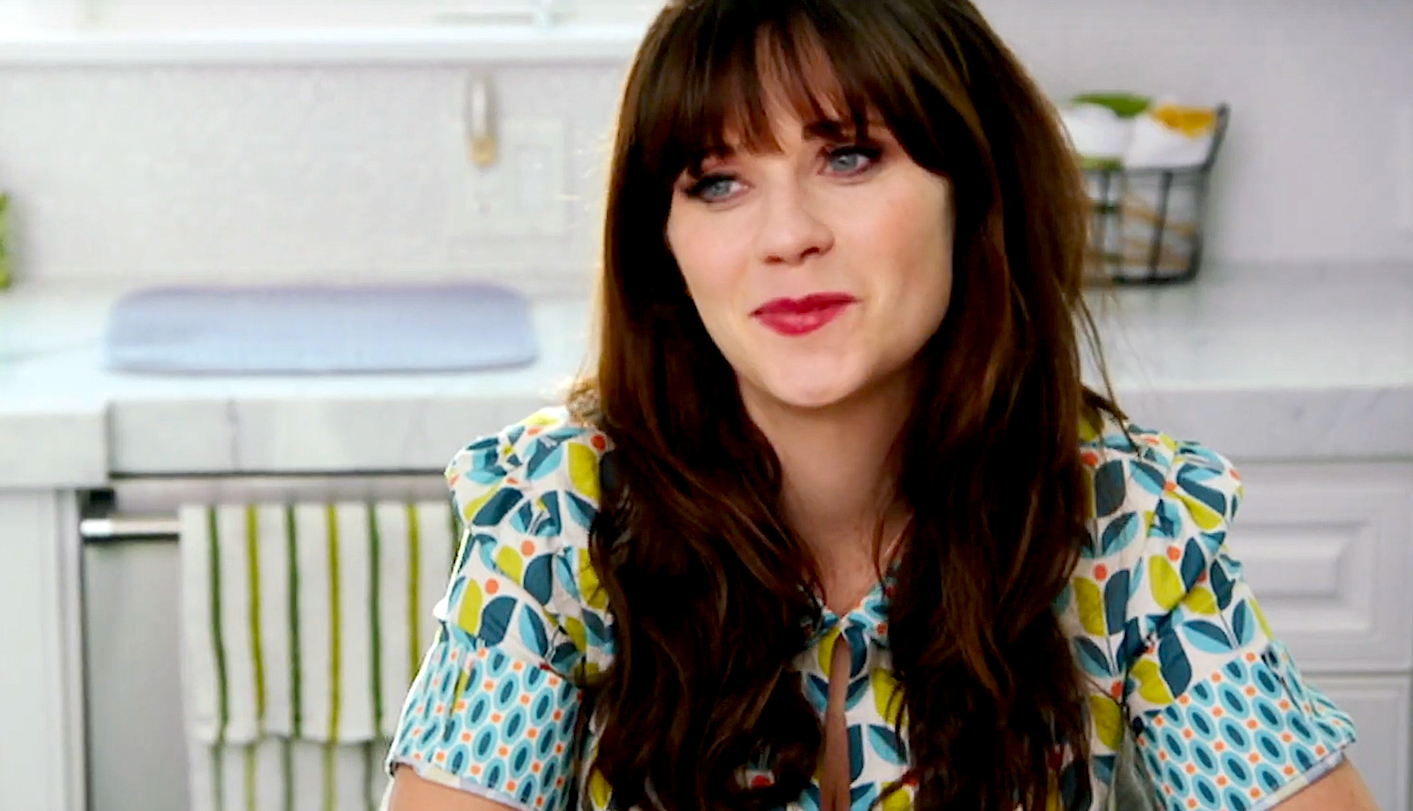 En person med langt hår og smell sitter på et kjøkken og har på seg en mønstret bluse med en slipsdetalj, og smiler litt