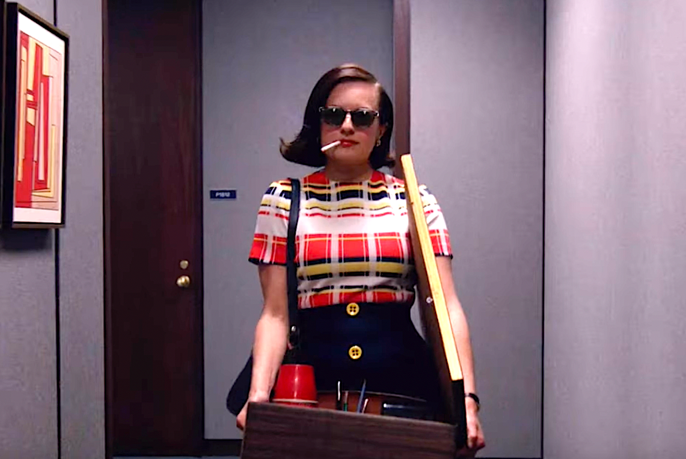 Person i retro plaid topp og solbriller holder en boks med kontorartikler, går trygt nedover en gang, og fremkaller en følelse av empowerment