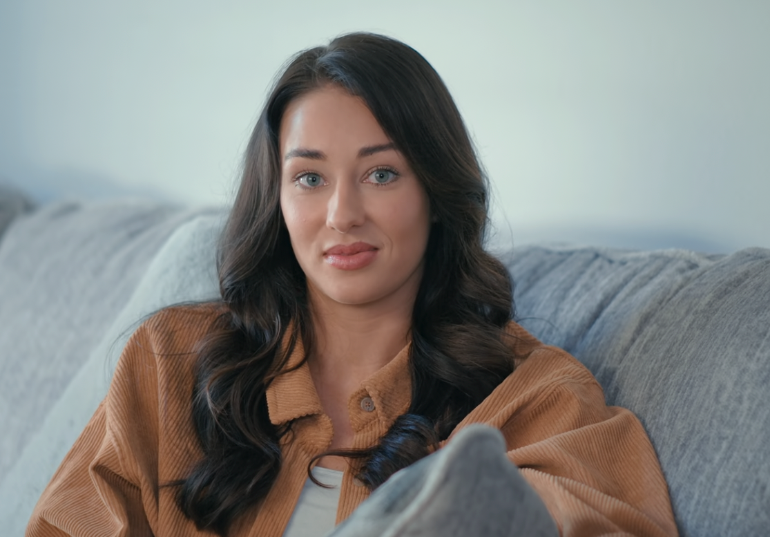 Person sitting on a couch wearing a casual jacket and top, with a neutral expression