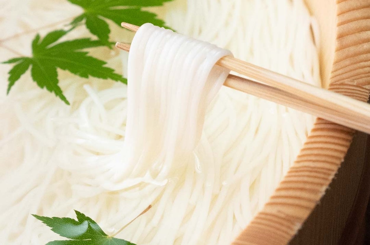 鉢に盛られた素麺を箸でつまんでいる様子。青い葉が水面に浮かんでいる。
