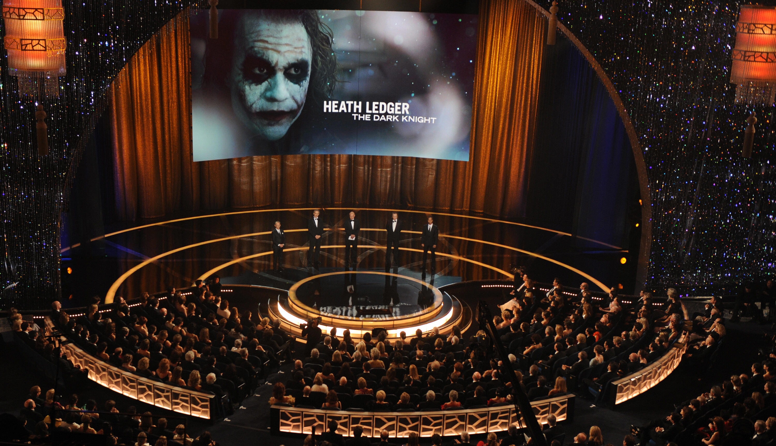 Audience at an award show watches a tribute to Heath Ledger, displayed on a large screen with his Joker image from "The Dark Knight."