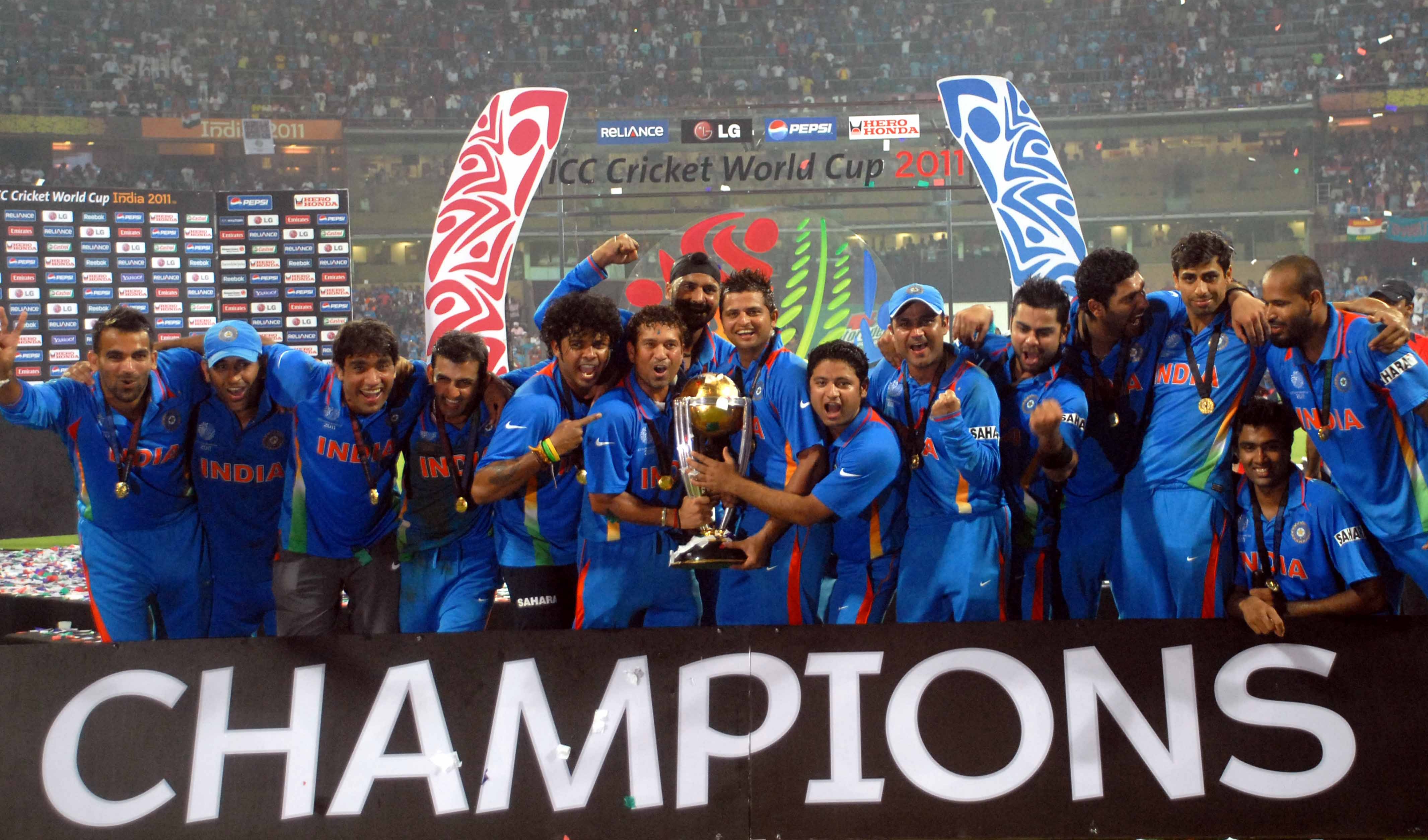 Cricket team celebrating with the World Cup trophy on stage, under a "Champions" banner, at a stadium event
