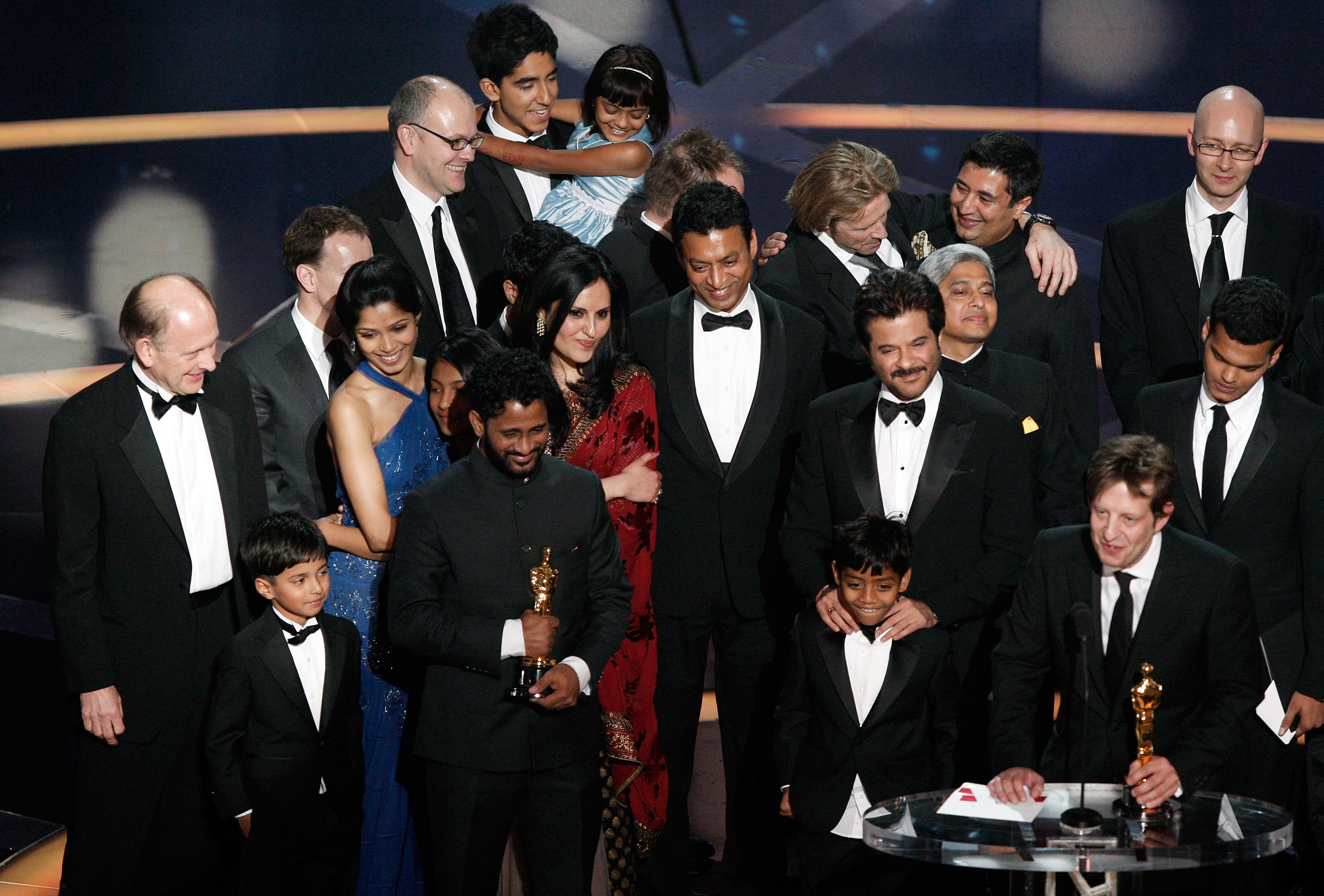 A large group of film cast and crew, dressed formally, celebrate on stage holding Oscars, showing joy and excitement