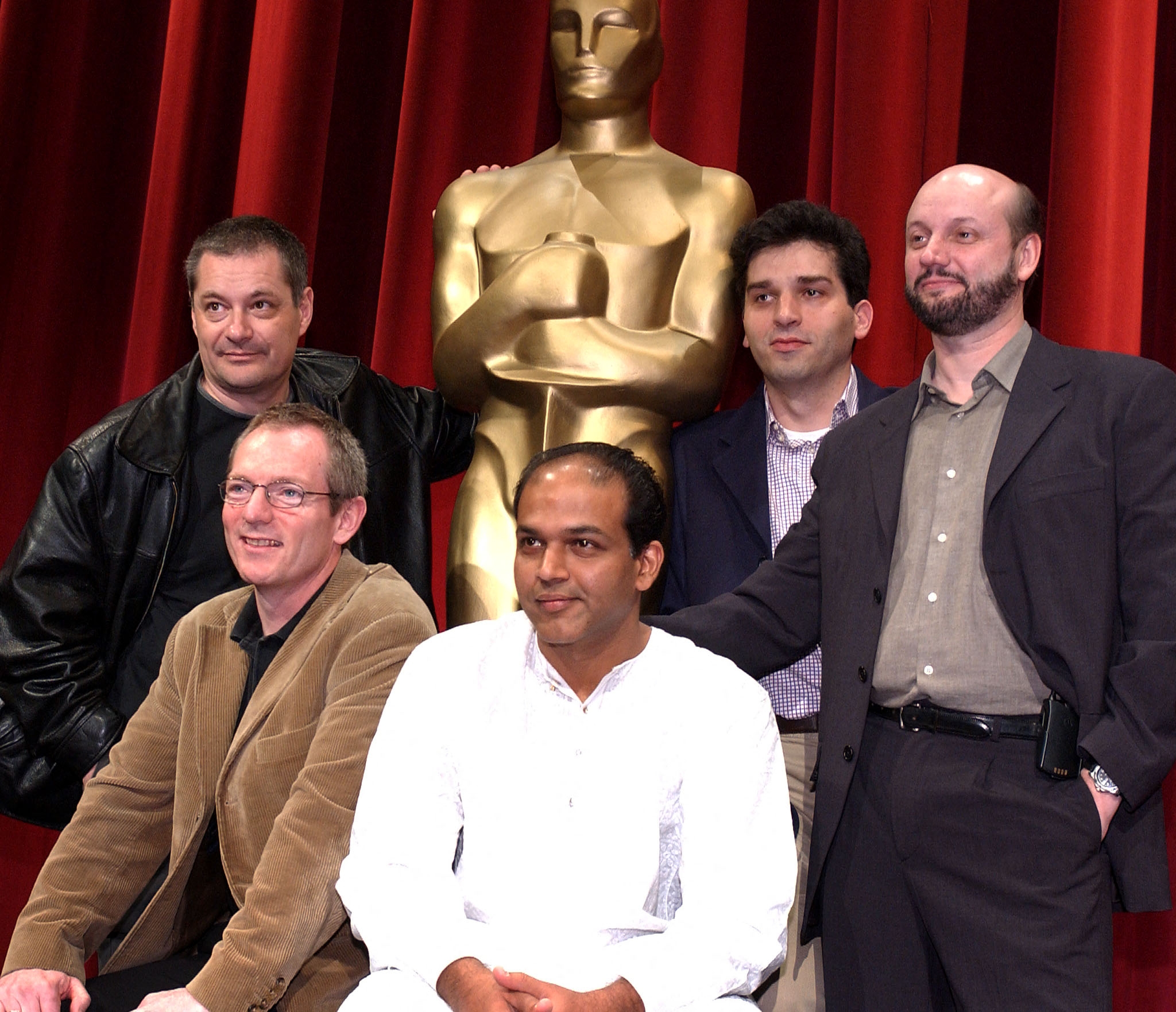 Five individuals pose in front of a large Oscar statue. Three are standing, two are seated. They wear casual and business attire