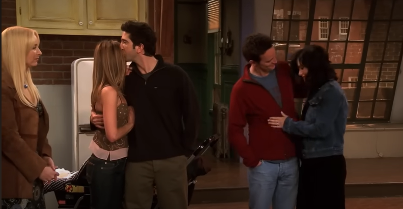 Five people from a sitcom stand in a living room. Two couples embrace, while another person watches with a neutral expression