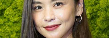 女性が白いシャツとジャケットを着て、フープイヤリングをつけて微笑んでいます。背景は緑の壁です。