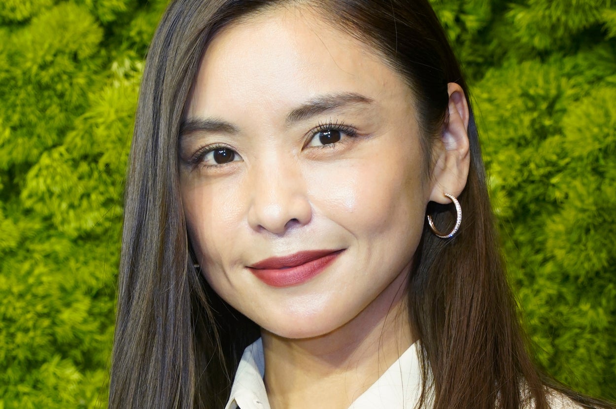 女性が白いシャツとジャケットを着て、フープイヤリングをつけて微笑んでいます。背景は緑の壁です。