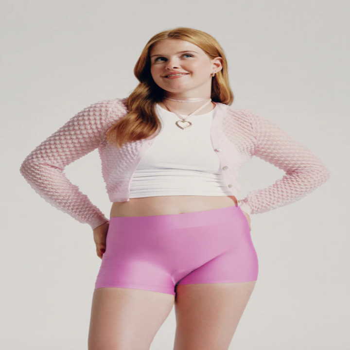 Person wearing a textured cardigan, white top, and pink shorts standing confidently with hands on hips, smiling upwards