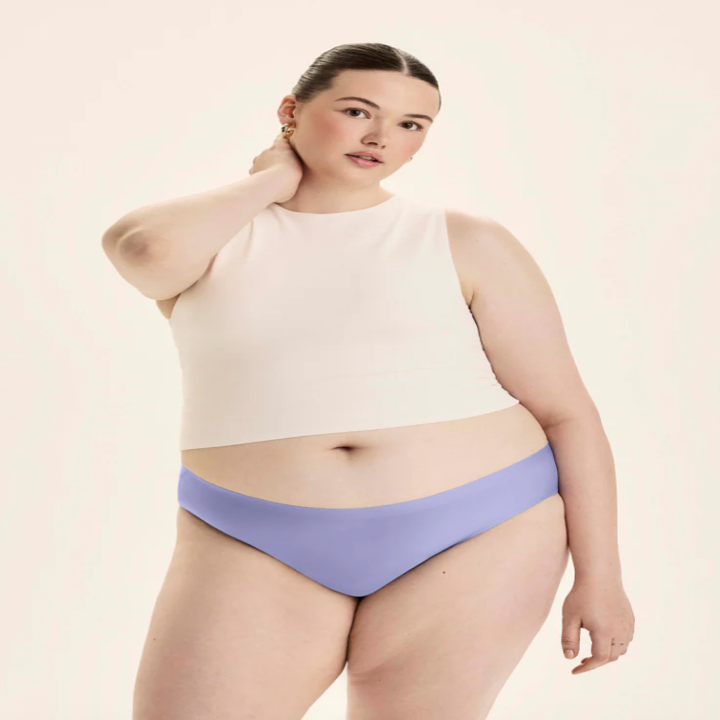 Model poses in a white sleeveless top and light underwear, looking confidently at the camera