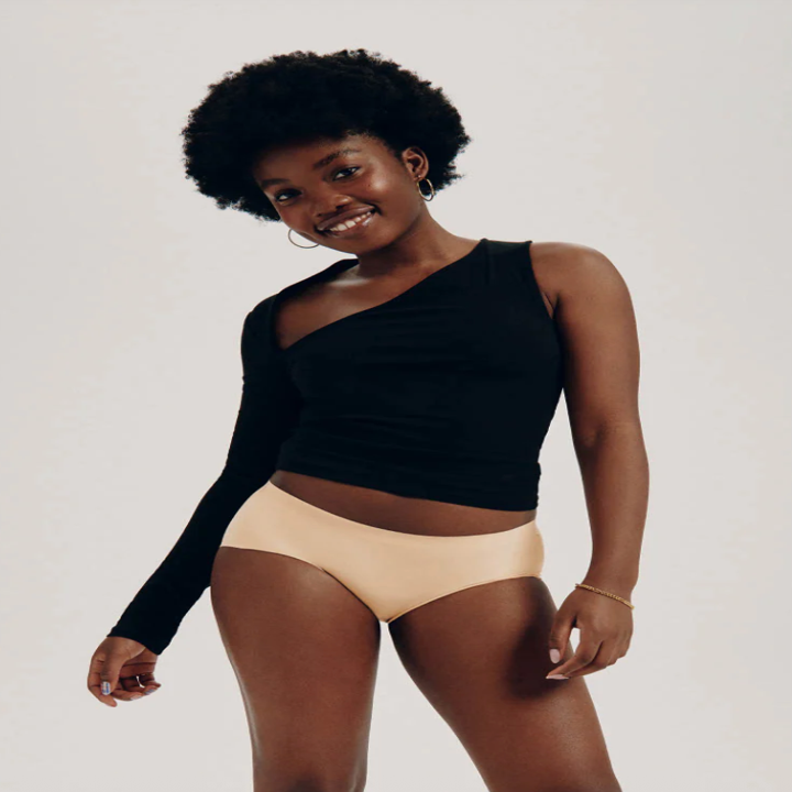 Person smiling, wearing a one-sleeve black top and beige shorts, posing confidently in a neutral studio setting