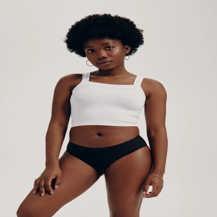 Person in a minimalist outfit featuring a white tank top and black briefs, standing confidently in a studio setting, suitable for shopping