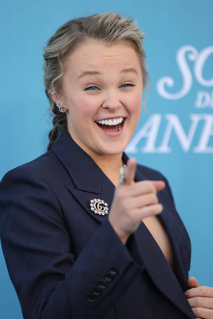 Person smiling and pointing, wearing a blazer with decorative pins, connected  a bluish  backdrop with indistinct text