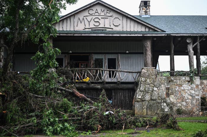 Tree branches and debris screen the structure of a rustic gathering with a "Camp Mystic" sign, perchance aft a storm