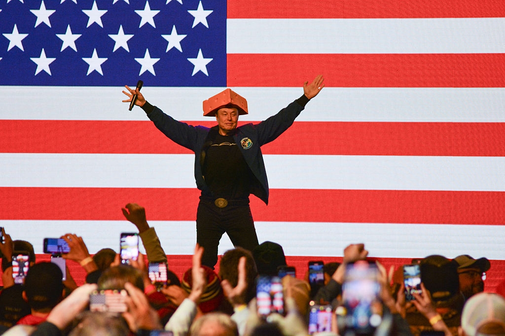 A man stands triumphantly on stage wearing a unique hat, holding a microphone, with an American flag backdrop. Audience members take photos
