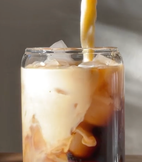 Iced coffee with milk being poured into a clear glass filled with ice, creating a swirling effect