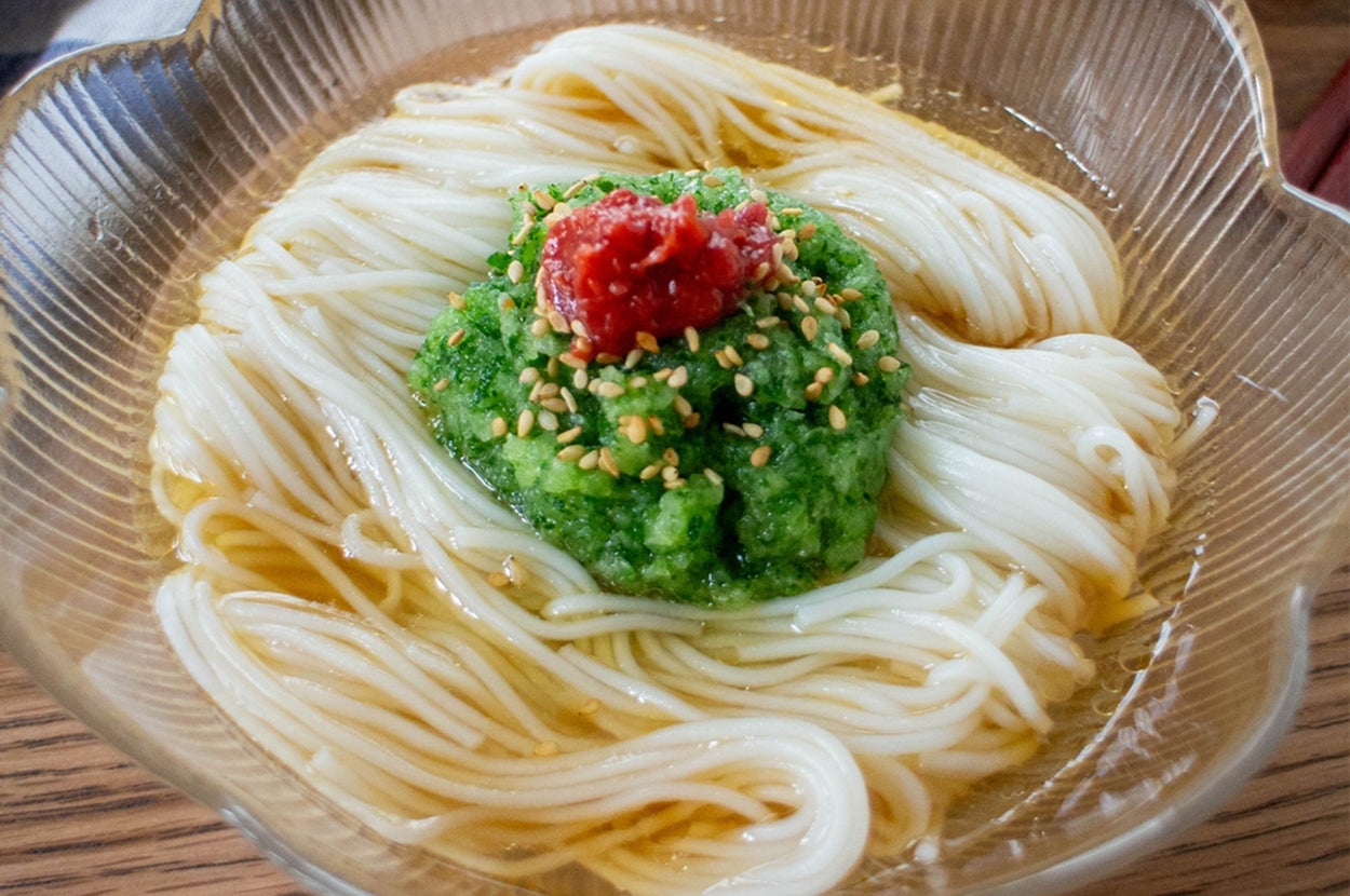 ガラスの器に盛られたそうめんの上に、緑色のソースと赤いトッピング、ゴマが飾られている。