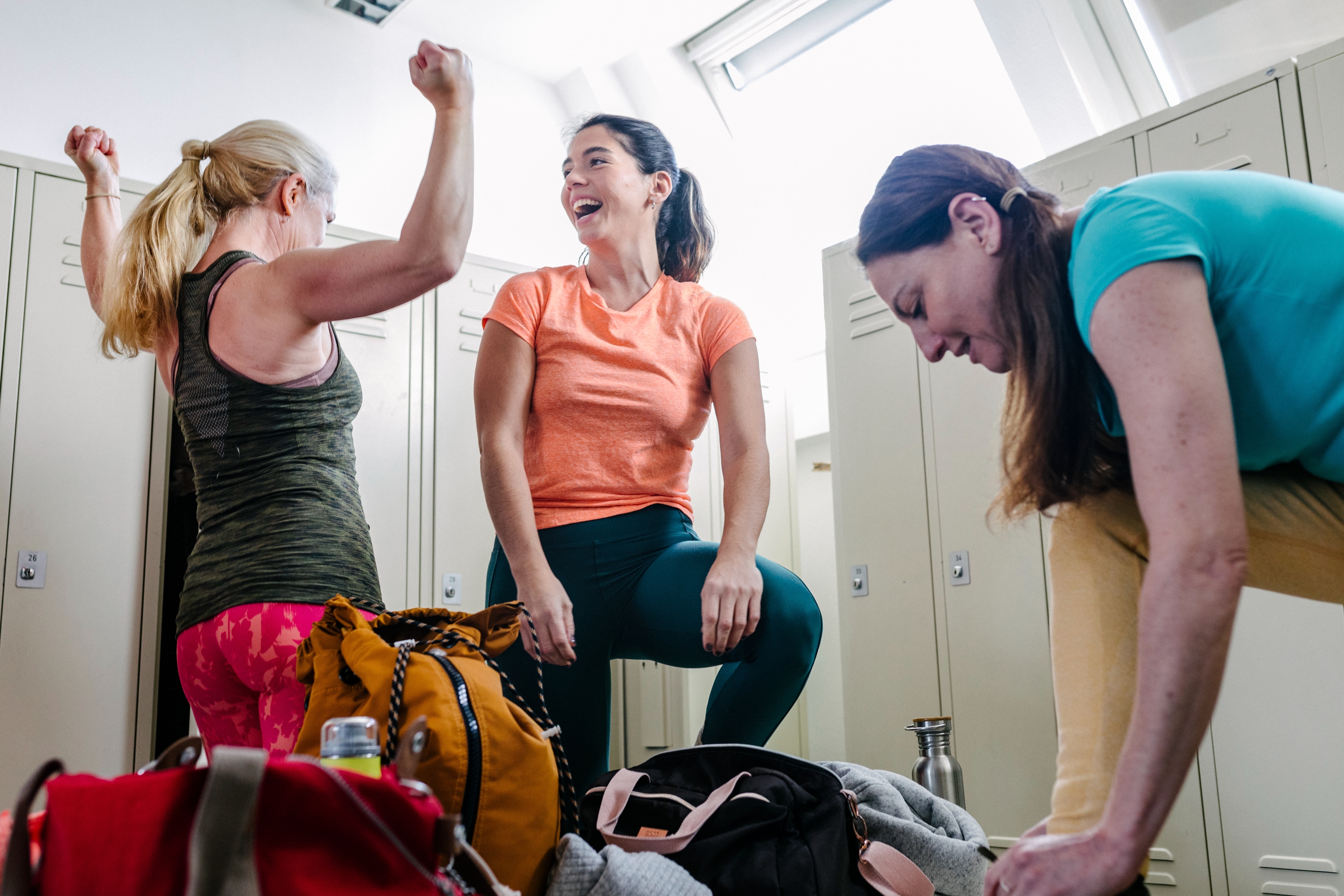 Three women in a locker room celebrate and laugh, with gym bags and lockers in the background. They appear energetic and cheerful