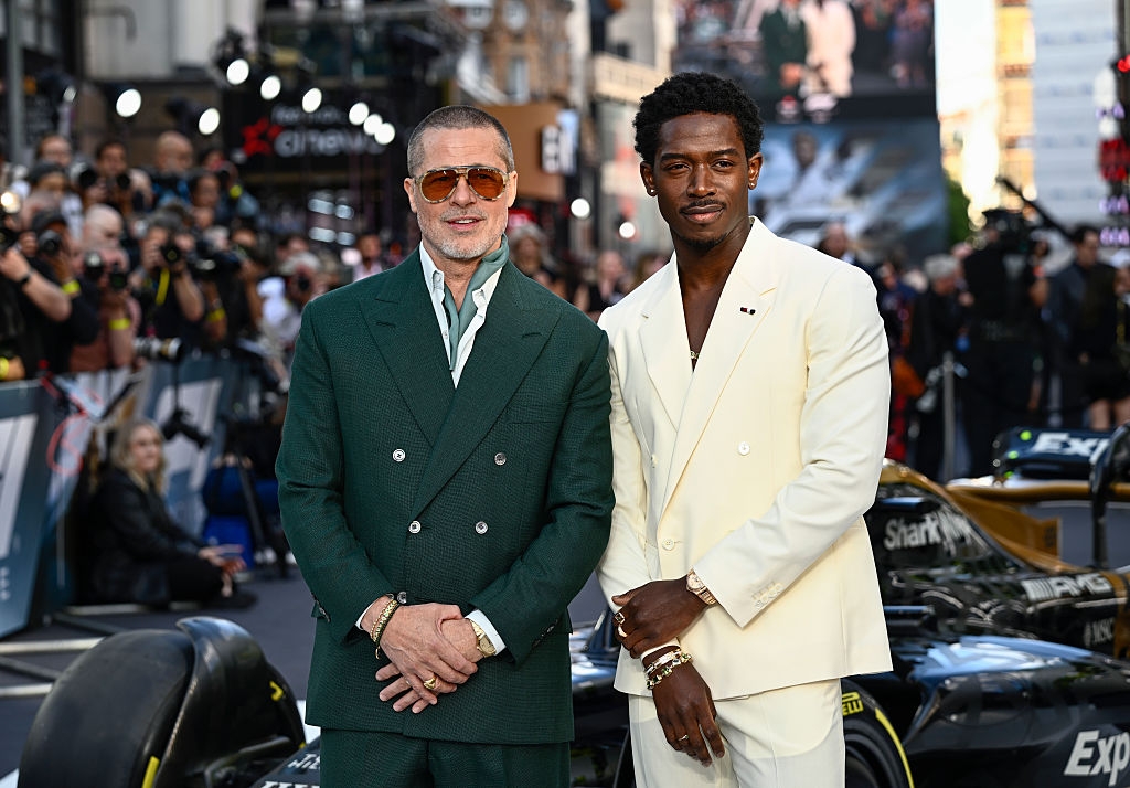 Two men on the red carpet, one in a green suit and sunglasses, the other in a white suit. They pose in front of a crowd with cameras in the background