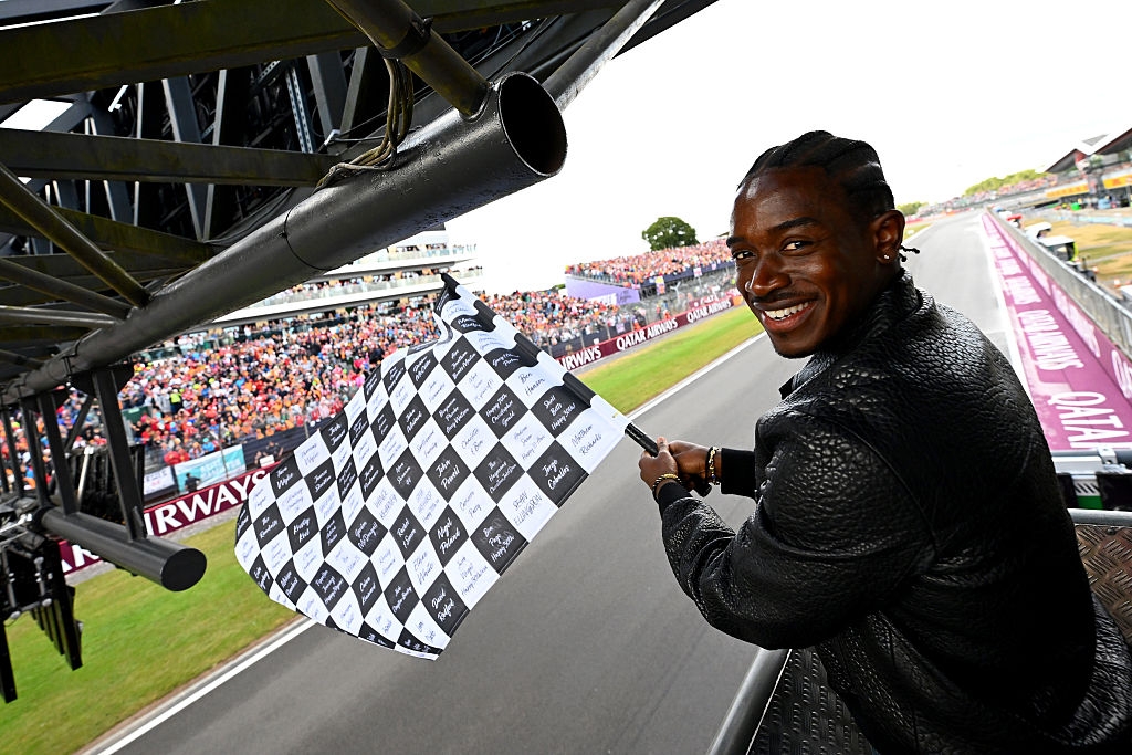 Person waving a black-and-white checkered flag at a racing event, smiling at the camera. Large crowd in the background