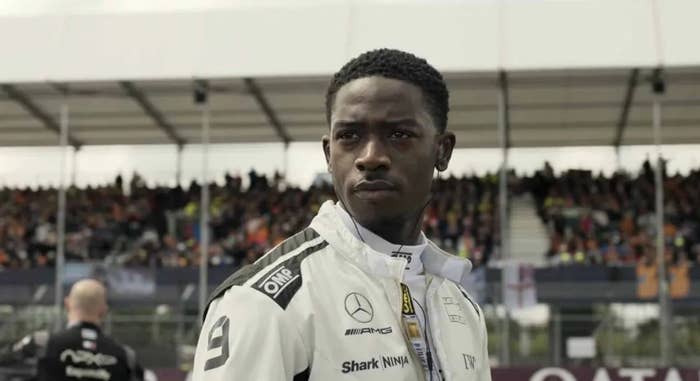 Person in racing suit stands on a race track, looking determined, with a crowded grandstand in the background