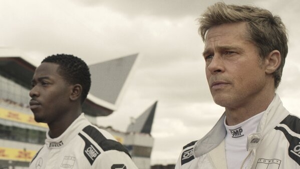 Two individuals in racing suits stand in front of a modern building, focused and determined