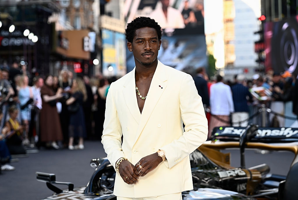 Man in a stylish suit poses confidently on a bustling street with a race car in the background, surrounded by people and cityscapes