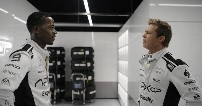 Two race car drivers in white racing suits face each other in a garage, engaged in conversation