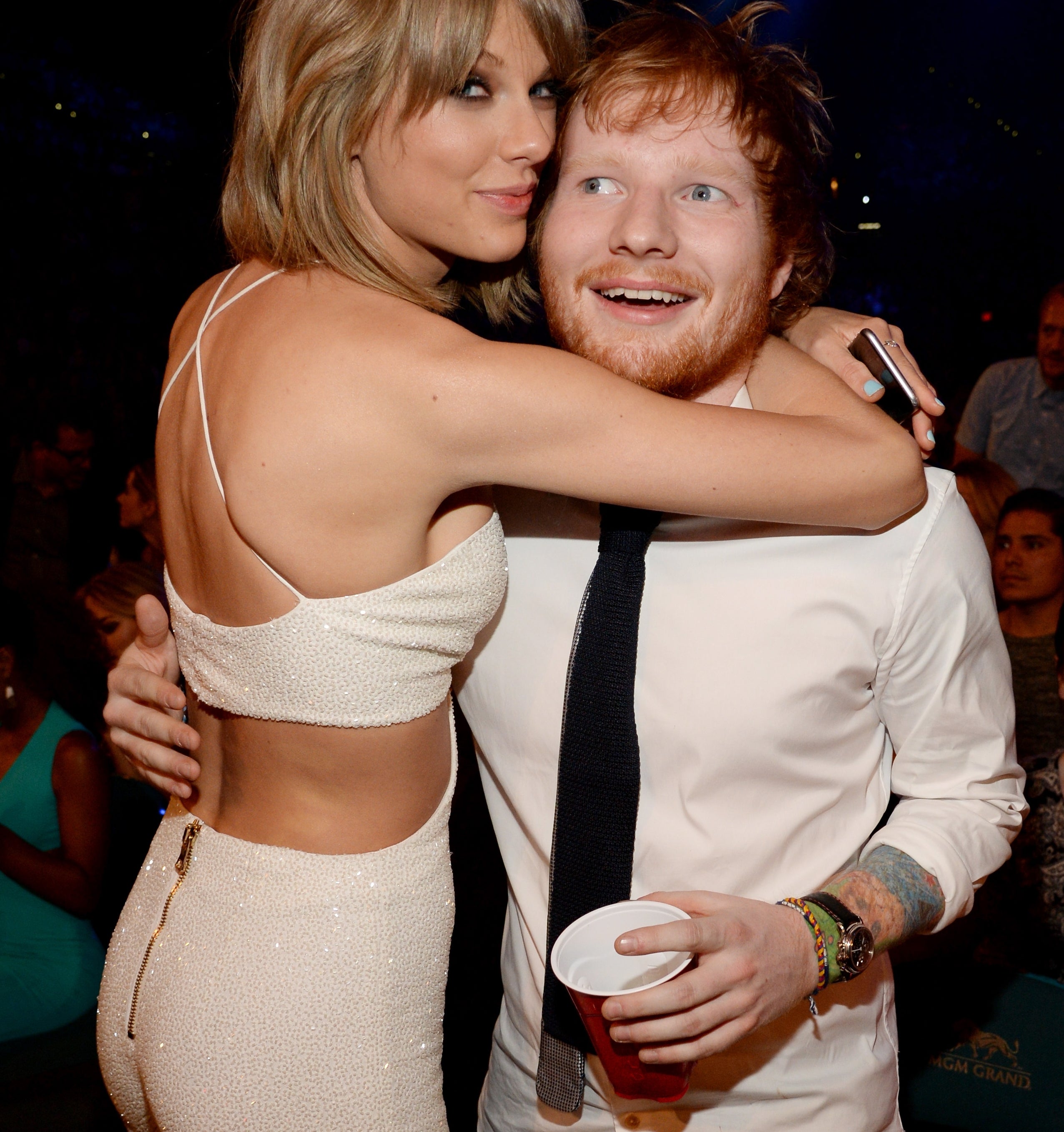 Two people smiling at an event. One is wearing a sparkling, sleeveless outfit, and the other is in a white shirt with a tie, holding a cup