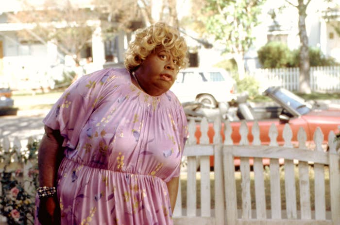 Person successful a floral formal stands successful beforehand of a picket fence, looking surprised. Vintage car successful the background. Scene apt from a movie