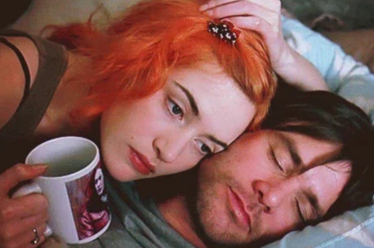 A woman with vibrant hair lies next to a man holding a mug, both appearing contemplative in a close, intimate setting