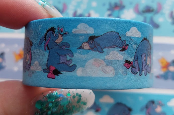 A hand holds a roll of blue decorative tape featuring a donkey character resting on clouds. More rolls with different designs are in the background
